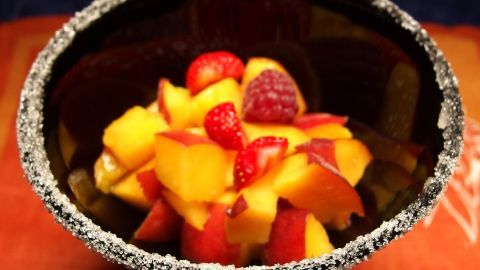 Ein Obstsalat liegt in einer Schale mit Zuckerrand, umgeben von bunten Früchten wie Pfirsichen und Himbeeren.