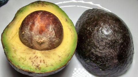 Eine halbierte, reife Avocado zeigt ihr saftiges Inneres neben einer ganzen, dunklen Avocado auf einem weißen Teller.