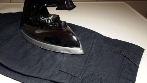 Ein schwarzer Bügel liegt auf einer schwarzen Hose, während er im Hintergrund auf einer hellen Arbeitsfläche steht.