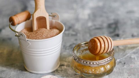 Ein kleiner Eimer mit braunem Zucker steht neben einem Honigglas und einem Holzlöffel, ideal für ein günstiges Lippenpeeling.