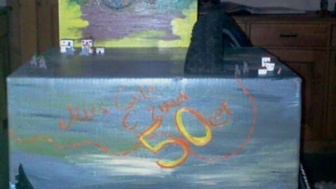 Zwei bemalte Geschenkboxen stehen zusammen in einem Raum, eine zeigt eine Landschaft mit Radfahrer, die andere feiert „50 Jahre“.
