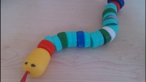 Eine Schlange aus PET-Flaschenverschlüssen liegt gekrümmt auf einem Tisch; bunte Verschlüsse bilden den Körper.
