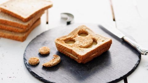 Ein Käse-Toast mit einem lächelnden Smiley-Gesicht liegt auf einer schwarzen Platte, umgeben von weiteren Brotscheiben.