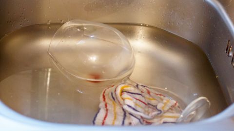 Gegen trübe Gläser: Gläser in Wasser mit einem Schuss Salmiakgeist geben, danach mit einem sauberen, trockenen Tuch polieren. Eine trübe Glas steht im Spülbecken, während ein Tuch im Wasser liegt, um es mit Salkmiakgeist zu reinigen.