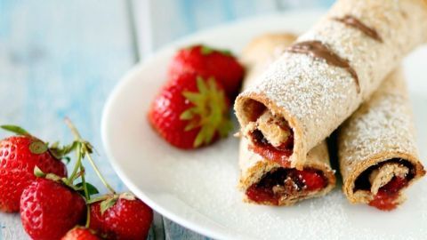 Cremige Erdbeer-Nutella-Toaströllchen liegen auf einem Teller, umgeben von frischen Erdbeeren, vor einem hellblauen Hintergrund.