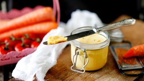 Eine zarte Tomaten-Möhren-Butter wird mit einem Messer aus einem Glas entnommen, umgeben von frischen Karotten und Tomaten.