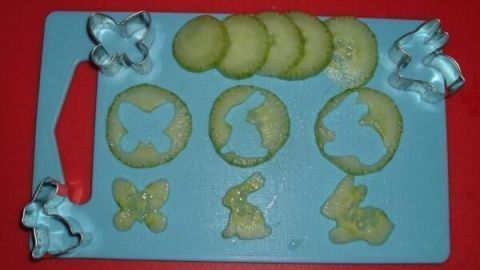 Gurkenscheiben in Form von Schmetterlingen und Hasen werden mit Ausstechformen auf einem blauen Brettchen präsentiert.