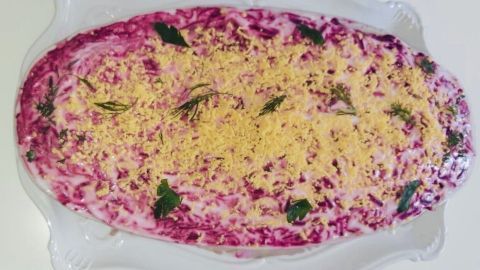 Ein Roter Heringssalat ist in einer ovalen Schüssel angerichtet, mit einer cremigen, lila-farbenen Oberfläche und überstreut mit gelbem, gemahlenem Ei. Die Salatoberfläche ist dekoriert mit frischen, grünen Blättern.