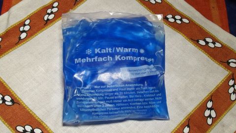 Ein Kühlelement in blauer Verpackung liegt auf einem gemusterten Tisch. Es ist zur Wärme- und Kältetherapie gedacht.