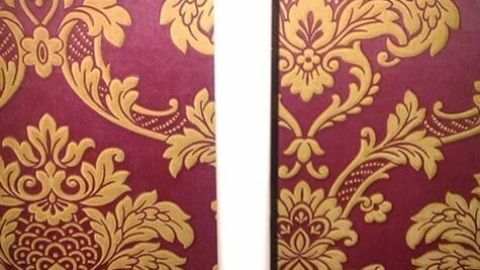 Ein langer, weißer Kerzenständer aus Gips hält eine brennende Kerze vor einer rot-goldenen floralen Tapete auf einem Tisch.
