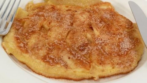 Pfannkuchen auf weißem Teller, bestreut mit Zucker, serviert mit Apfelmus, liegt neben Messer und Gabel auf einem Tisch.