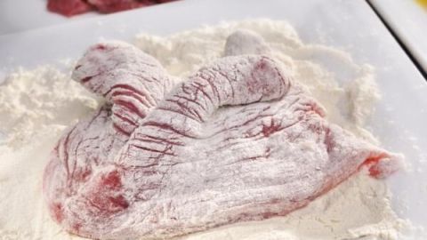 Ein herzförmiges Stück Fleisch liegt in Mehl auf einem Teller, bereit zum Panieren. Im Hintergrund mehr rohes Fleisch.