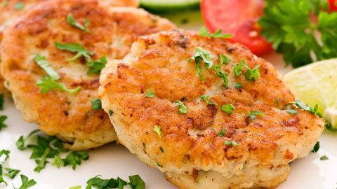 Drei goldbraune Räucherfisch-Frikadellen liegen auf einem Teller, garniert mit frischem Grün und umgeben von Tomate und Limette.