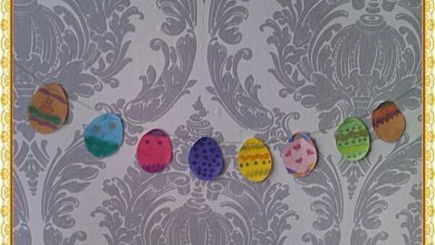 Papier-Ostereier hängen als Girlande vor einer grauen, verzierten Tapete und zeigen bunte Muster.