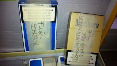 Zwei Schachteln beschriftet mit „FLUR: SCHRAUB“ und „WOHNZIMMERSCHRANK-SCHUBLADE TINE“ liegen in einem Karton.