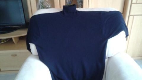Ein Cashmere Pullover ist über einen hellen Sessel gelegt, im Hintergrund steht ein Holzschrank mit Dekorationen.