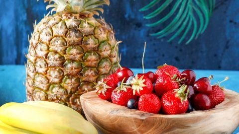 Eine frische Ananas steht neben einer Holzschüssel voller Beeren, umgeben von Bananen, Tomaten und einer Gurke.