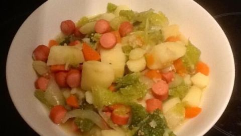 Eine weiße Schale mit Wirsingeintopf, bestehend aus Kartoffeln, Karotten und Wiener Würstchen, auf einem dunklen Untergrund.