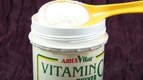 Ein gelber Löffel hebt weißen Vitamin C-Pulver aus einem Behälter, auf einem dunklen Hintergrund.