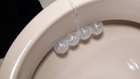 Ein durchsichtiger Duftspender hängt am Rand einer Toilette, während der Badezimmerboden in sanften Farbtönen gehalten ist.