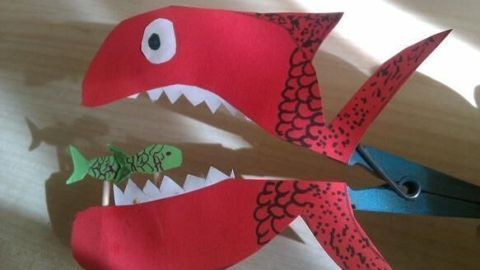 Roter Papierfisch mit Zähnen aus Pappe auf Wäscheklammer schnappt nach kleinem grünen Fisch; Schattenwurf auf Holztisch.