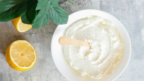 Eine cremige Gesichtsmaske in einer Schüssel wird mit einem Holzspatel verrührt, um samtweiche Haut zu erzielen. Neben der Schüssel liegen eine halbe Zitrone und grüne Blätter.