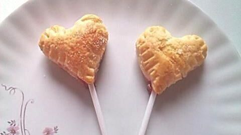 Zwei herzförmige Blätterteig-Snacks auf einem weißen Teller mit Blumenmuster stehen aufrecht auf Holzstäbchen.