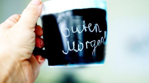 Eine Tasse mit Tafellack wird hochgehalten, auf deren Oberfläche "Guten Morgen!" steht, vor unscharfem Hintergrund.
