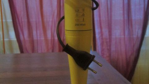 Ein gelber Stabmixer steht auf einem Tisch, das Kabel ist um ihn gewickelt, in einem Raum mit Vorhängen.