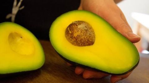 Eine Hand hält eine halbierte Avocado mit sichtbarem Avocadokern, auf einem Holztisch liegend.