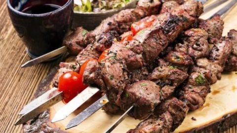 Souvlaki-Spieße, frisch gegrillt mit Tomaten, liegen auf einem Holzbrett; daneben steht ein Becher Rotwein und Salat.