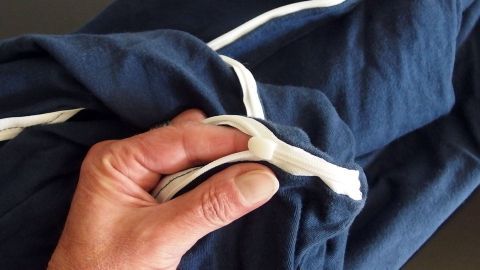 Eine Hand hält einen defekten Reißverschluss an einer blauen Bettwäsche, die verdreht auf einem Tisch liegt.