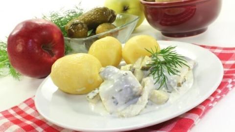 Heringsstipp mit Sahne und Dill auf Teller, begleitet von Kartoffeln. Im Hintergrund Äpfel und Gurken auf kariertem Tuch.