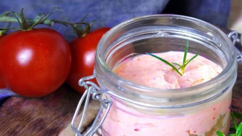 Ein Glas Pesto Rosso steht auf einem Holzbrett, umgeben von roten Tomaten und frischen Kräutern in einer rustikalen Umgebung.