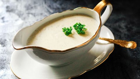 Eine elegante Sauciere mit weißer Milchsoße, garniert mit frischer Petersilie, steht vor einem dunklen Hintergrund.