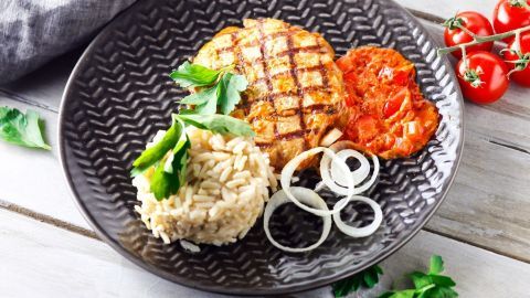 Ein schmackhaftes Gericht besteht aus einer gegrillten Patty, Paprika-Zwiebelsoße, Reis und frischen Kräutern auf einem dunklen Teller.