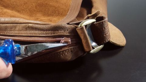 Eine Hand hält ein Werkzeug, um einen Reißverschluss einer braunen Tasche zu reparieren, der nicht mehr richtig schließt.