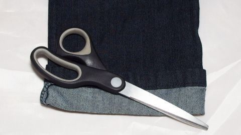 Eine Schere liegt neben dem Originalsaum einer dunkelblauen Jeans, bereit, um diese zu kürzen.