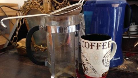 Eine French Press steht auf einem Tisch, während heißer Kaffee in eine Tasse mit dem Schriftzug „COFFEE“ gegossen wird.
