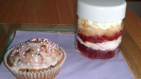Ein Muffin mit Zuckerguss und Streuseln liegt auf einem lila Serviettenuntergrund neben einem geschichteten Torte im Glas.