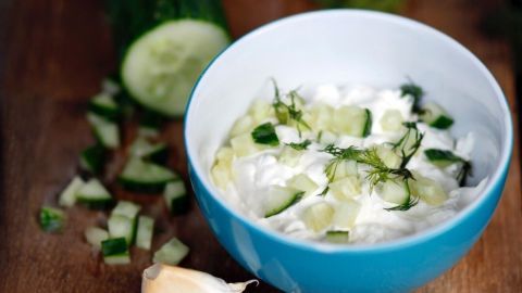 Eine Schüssel mit Tzatziki aus Quark und Gurkenstückchen steht auf einem Holzbrett, umgeben von gehacktem Knoblauch.