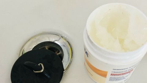 Ein Badewannenstöpsel wird gehandhabt, während eine Creme daneben liegt; der Stöpsel verhindert das Ablaufen des Wassers.