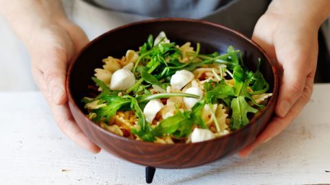Eine Person hält eine Schüssel Nudelsalat mit Rucola und Mozzarella, umgeben von einem hellen, rustikalen Tisch.