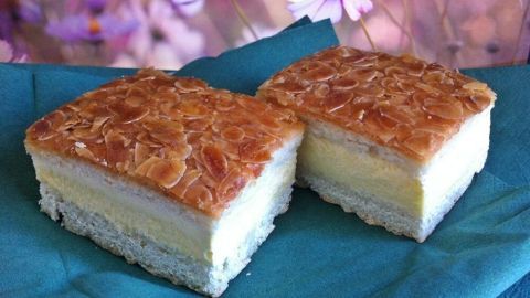 Ein Bienenstich, ein saftiges Stück Kuchen mit einer goldbraunen Mandelschicht, liegt auf einem grünen Tuch vor einer blühenden Hintergrundszenerie.