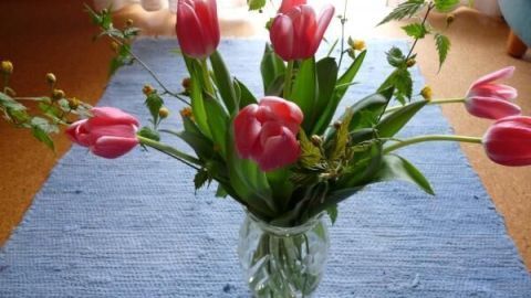 Ein bunter Blumenstrauß mit rosa Tulpen steht in einer Glasvase auf einem blauen Teppich im hellen Raum.