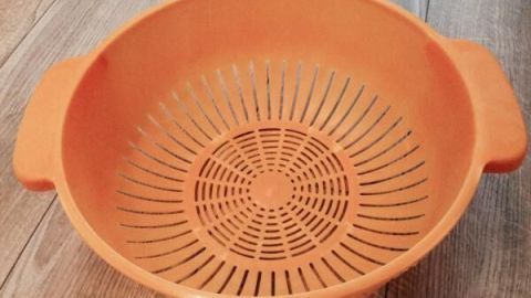 Ein orangefarbener Pflanzkorb liegt auf einem Holzboden. Der Korb hat ein Gittermuster und breite Griffe.