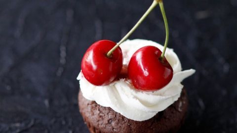 Eine schokoladige Muffin mit einer Buttercremehaube, gekrönt von zwei leuchtend roten Kirschen, auf einem dunklen Untergrund.