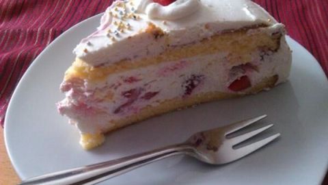 Ein Stück Erdbeertorte mit Sahne und Erdbeerstücken liegt auf einem weißen Teller, eine Gabel daneben.