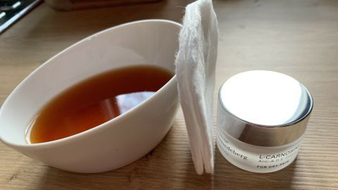 Eine Schale mit braunem Flüssigkeit steht neben einer Creme und Wattepads auf einem Holzuntergrund, um trockene, schuppige Haut zu behandeln.