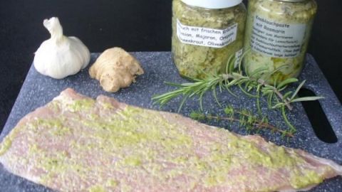 Eine Fischfilet mit würziger Kräuterpaste liegt auf einem Schneidebrett, umgeben von Knoblauch, Ingwer und Gläsern mit Kräutermischung.